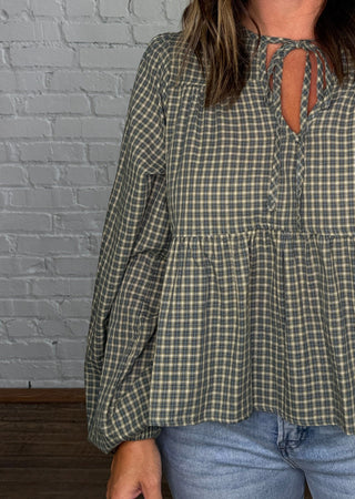 A woman wearing a green and beige gingham long-sleeve blouse with a tie-neck detail, paired with light-wash jeans, standing in front of a white brick wall.