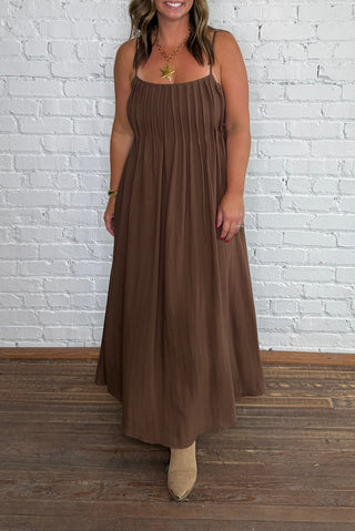 A woman wearing a sleeveless brown midi dress with a pleated bodice and flowing skirt, paired with tan ankle boots.