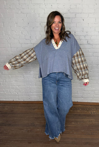 A woman modeling a mineral washed denim top with plaid sleeves & floral hem paired wide-leg blue jeans, standing against a white painted brick wall on a wooden floor. 