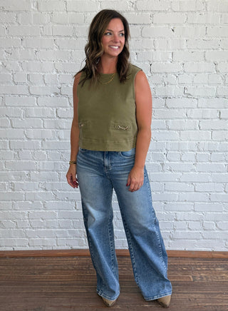 A woman stands in a room with a white brick wall and dark wood floor, wearing a sleeveless olive green top with gold equestrian horse bit faux pocket detail and wide-leg blue jeans.
