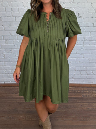 A woman modeling an olive green short-sleeve dress with a buttoned split neckline, standing against a white brick wall and wood floor.
