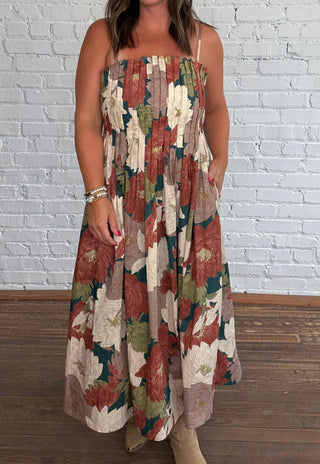 A woman wearing a spaghetti strap dress with a multicolor floral pattern in earthy tones plus pockets, standing on a dark wood floor against a white brick wall.