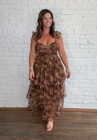 A woman wearing a brown tiered tulle maxi dress with ruffled shoulder straps, standing on a dark wood floor against a white painted brick wall.