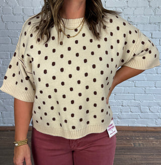 A woman stands in a studio with a white brick wall and wood floor, wearing a cream knit sweater with brown polka dots and dusty rose pants.