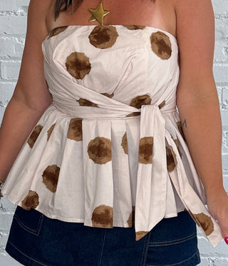 A woman modeling a cream strapless top with brown polka dots, dark denim short, and brown leather cowboy boots, standing against a white brick wall.