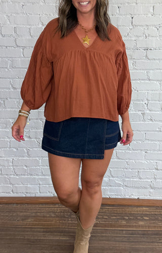 A woman modeling a rust-colored long-sleeve blouse with a V-neck and gathered shoulders, paired with a dark denim mini skort and beige ankle boots, standing against a white painted brick wall on a wooden floor.