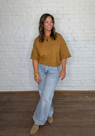 A woman stands in front of a white brick wall wearing a mustard brown short-sleeve knit top, light blue jeans, and tan ankle boots.