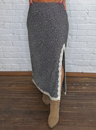 A woman wearing a brown and blue floral midi skirt with cream lace trim, standing against a white brick wall and wooden floor.