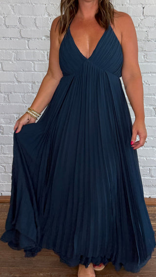 A woman modeling a deep V-neck, floor-length pleated maxi dress in dark teal blue, standing against a white painted brick wall on a wooden floor.