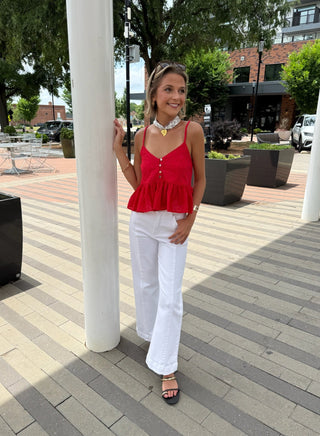 Woman in a red top and white pants standing on a wooden deck with trees and buildings in the background.