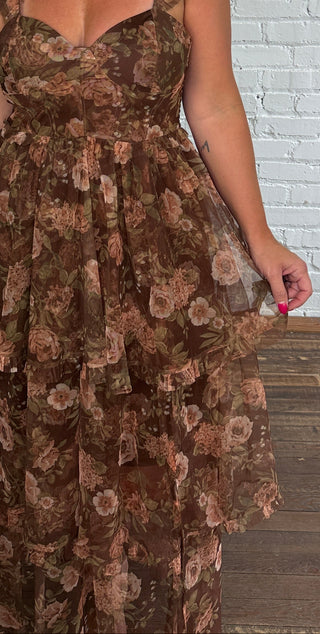 A woman wearing a brown floral tiered maxi dress with ruffled shoulder straps, standing in front of a white brick wall and dark wood floor.