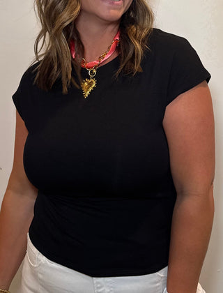 Woman wearing a black top and white pants standing against a plain wall.