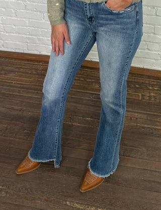 Woman wearing a green patterned shirt and blue jeans standing against a white brick wall.