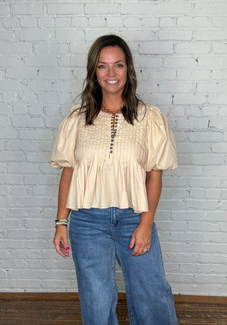 A woman modeling a cream-colored short-sleeve blouse with puff sleeves and a button-up front, paired with wide-leg blue jeans, standing against a white brick wall and wooden floor.