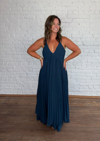 A woman modeling a deep V-neck, floor-length pleated maxi dress in dark teal blue, standing against a white painted brick wall on a wooden floor.