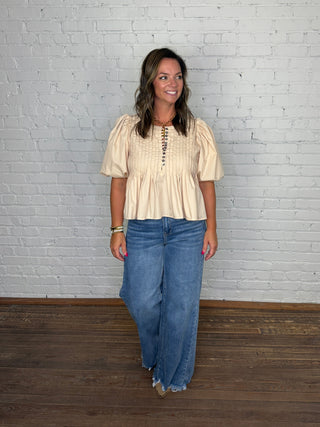 A woman modeling a cream-colored short-sleeve blouse with puff sleeves and a button-up front, paired with wide-leg blue jeans, standing against a white brick wall and wooden floor.