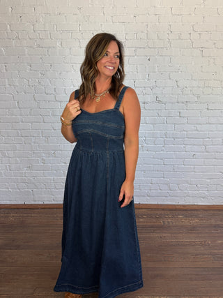 A woman modeling a dark blue denim maxi dress with a smocked bodice, standing against a white brick wall and wooden floor.