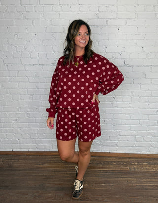 A woman modeling a matching burgundy and pink polka dot two-piece set standing against a white brick wall and wooden floor.