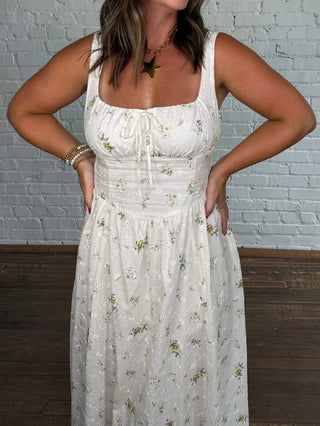 A woman modeling a spaghetti strap cream maxi dress with a delicate floral pattern, standing against a white brick wall on a dark wood floor.