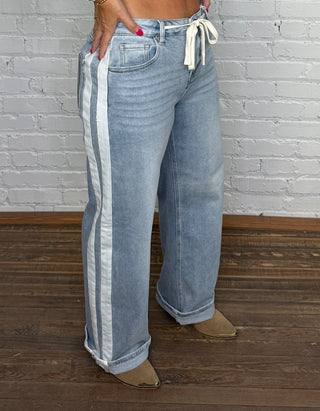 A person wearing light-wash wide-leg jeans with a white side stripes and white drawstring tie at the waist, standing on a dark wood floor against a white brick wall.