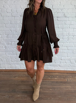 A woman modeling a dark brown long-sleeve mini dress with a button-up front and tie waist, paired with beige cowboy boots, standing in front of a white brick wall on a wooden floor.