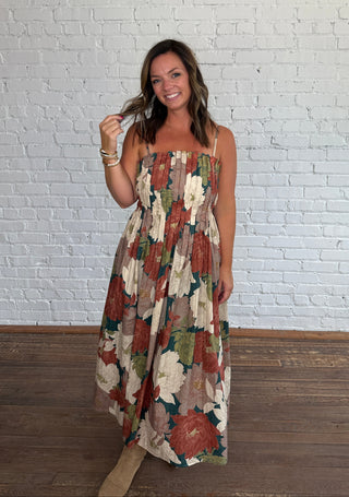 A woman wearing a spaghetti strap dress with a multicolor floral pattern in earthy tones, standing on a dark wood floor against a white brick wall.