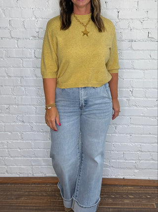 A woman stands against a white brick wall wearing a mustard yellow knit top, light blue wide-leg jeans, and tan boots.