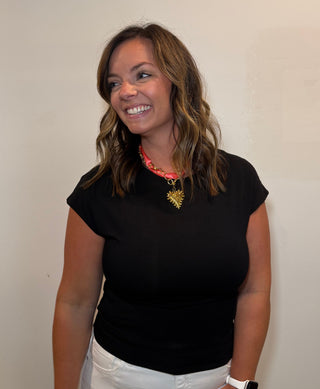 Woman wearing a black top and white pants standing against a plain wall.