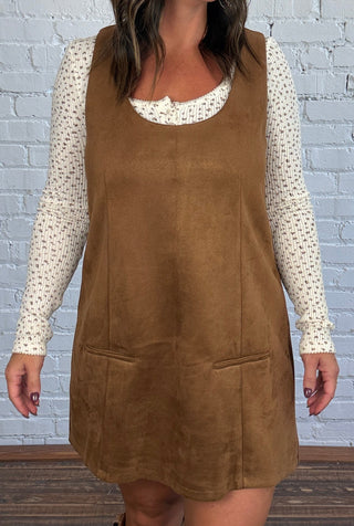 A woman modeling a brown dress over a cream, floral, long-sleeve top, paired with brown cowboy boots, standing against a white brick wall.