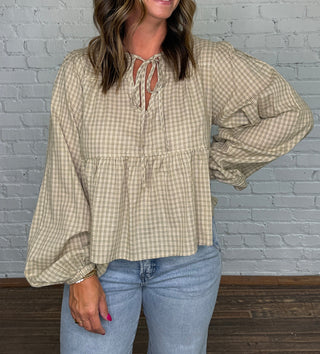 A woman wearing a beige and green gingham long-sleeve blouse with a tie-neck detail, paired with light blue jeans, standing in front of a white brick wall.