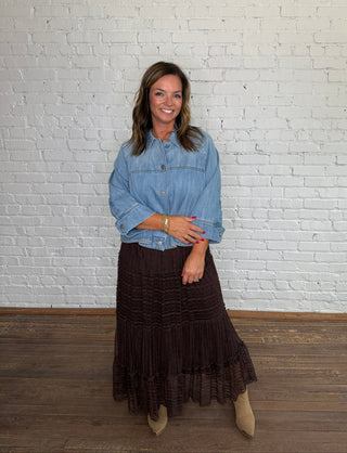 A woman stands in front of a white brick wall wearing a cropped denim top and a dark brown tiered lace midi skirt, paired with taupe ankle boots.