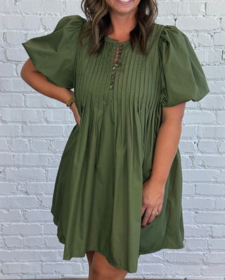 A woman modeling an olive green puff-sleeve mini dress with a gathered smocked bodice and front button placket, paired with tan suede ankle boots, standing against a white brick wall and dark wood floor.
