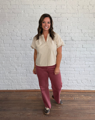 A woman modeling a cream and beige vertically striped short-sleeve blouse with puff sleeves and v-neck neckline, paired with dusty rose pants and patterned sneakers, standing against a white brick wall and dark wood floor.