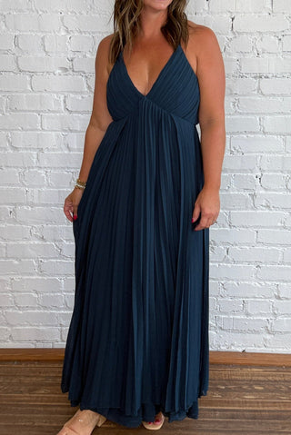 A woman modeling a deep V-neck, floor-length pleated maxi dress in dark teal blue, standing against a white painted brick wall on a wooden floor.