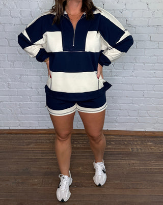 A woman modeling a matching navy blue and cream athletic set consisting of a half-zip long-sleeve top and shorts, standing against a white brick wall and wooden floor.