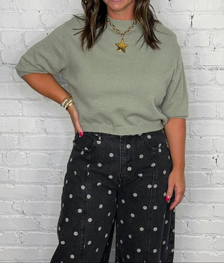 A woman stands against a white brick wall wearing a sage green short-sleeve t-shirt and black barrel jeans with white polka dots, paired with tan boots.