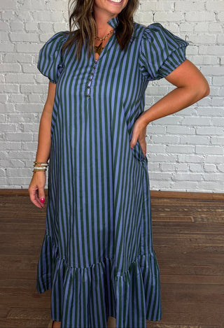 A woman wearing a blue and green vertically striped midi dress with puff sleeves and beige ankle boots, standing against a white brick wall on a wooden floor.