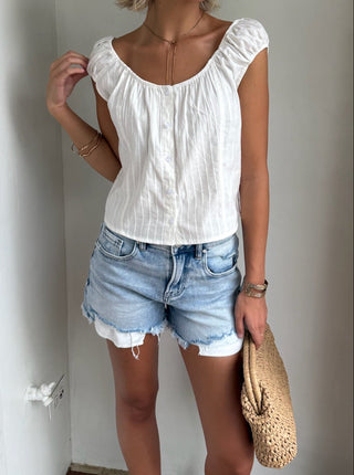 Woman wearing a white top and denim shorts standing against a plain wall.