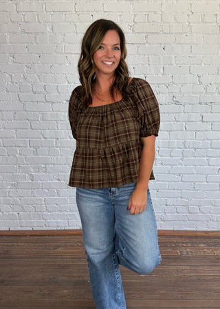 A woman modeling a brown, tan and taupe plaid puff-sleeve top with light-wash blue jeans, standing in front of a white brick wall and wooden floor.