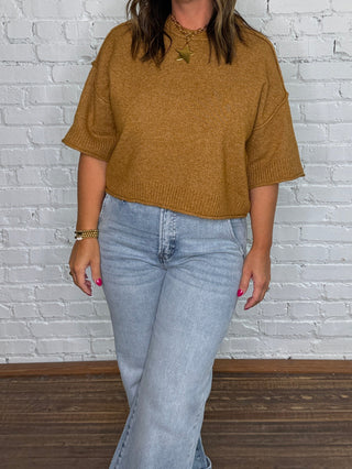 A woman stands in front of a white brick wall wearing a mustard brown short-sleeve top, light blue jeans, and tan boots.