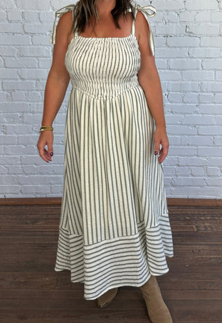 A woman modeling a sleeveless, smocked maxi dress with vertical cream and olive green stripes, featuring tie straps on the shoulders and a tiered skirt, standing against a white brick wall and dark wood floor.