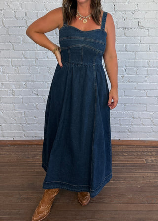 A woman modeling a dark blue denim maxi dress with a smocked bodice and thin straps, standing against a white painted brick wall on a wooden floor.