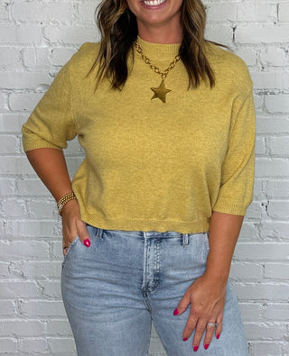 A woman stands in front of a white brick wall wearing a mustard yellow knit top, light-wash wide-leg jeans, and tan ankle boots.