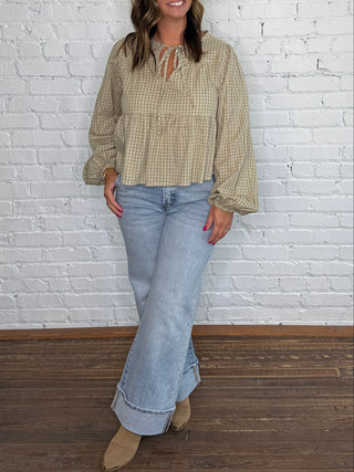 A woman stands against a white brick wall on a dark wood floor, wearing a beige and green gingham long-sleeve blouse with a tie neckline, light blue flared jeans, and tan boots.
