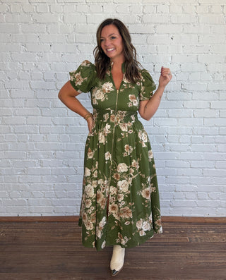 A woman modeling a green floral midi dress with puff sleeves and a front zipper, standing against a white brick wall on a wooden floor.