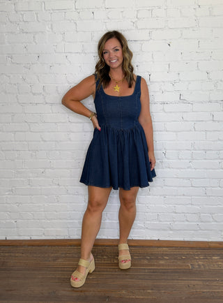 A woman modeling a sleeveless dark blue denim mini dress with a gathered waist.