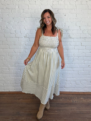 A woman modeling a sleeveless, square-neck midi dress with thin vertical stripes, tied shoulder straps, and a smocked waist, standing in front of a white brick wall on a wooden floor.
