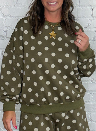 A woman modeling an olive green and white polka dot lounge set consisting of a long-sleeve sweatshirt and matching shorts, standing against a white brick wall and wooden floor.