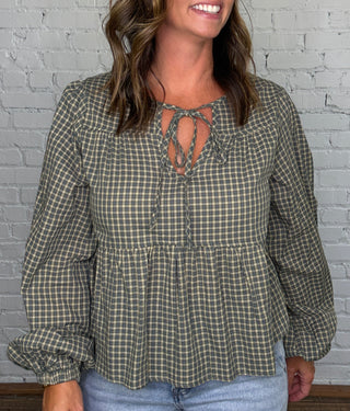 A woman modeling a long-sleeve green and beige gingham blouse with a tie-front neckline, paired with light-wash jeans, standing against a white brick wall and dark wood floor.