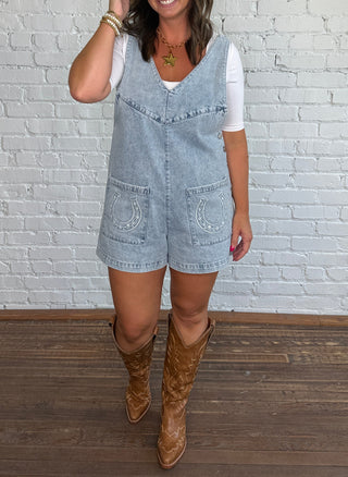 A woman stands in front of a white brick wall wearing a short, light blue denim overall romper with a horseshoe embroidered on each pocket, a white t-shirt underneath, and brown cowboy boots.